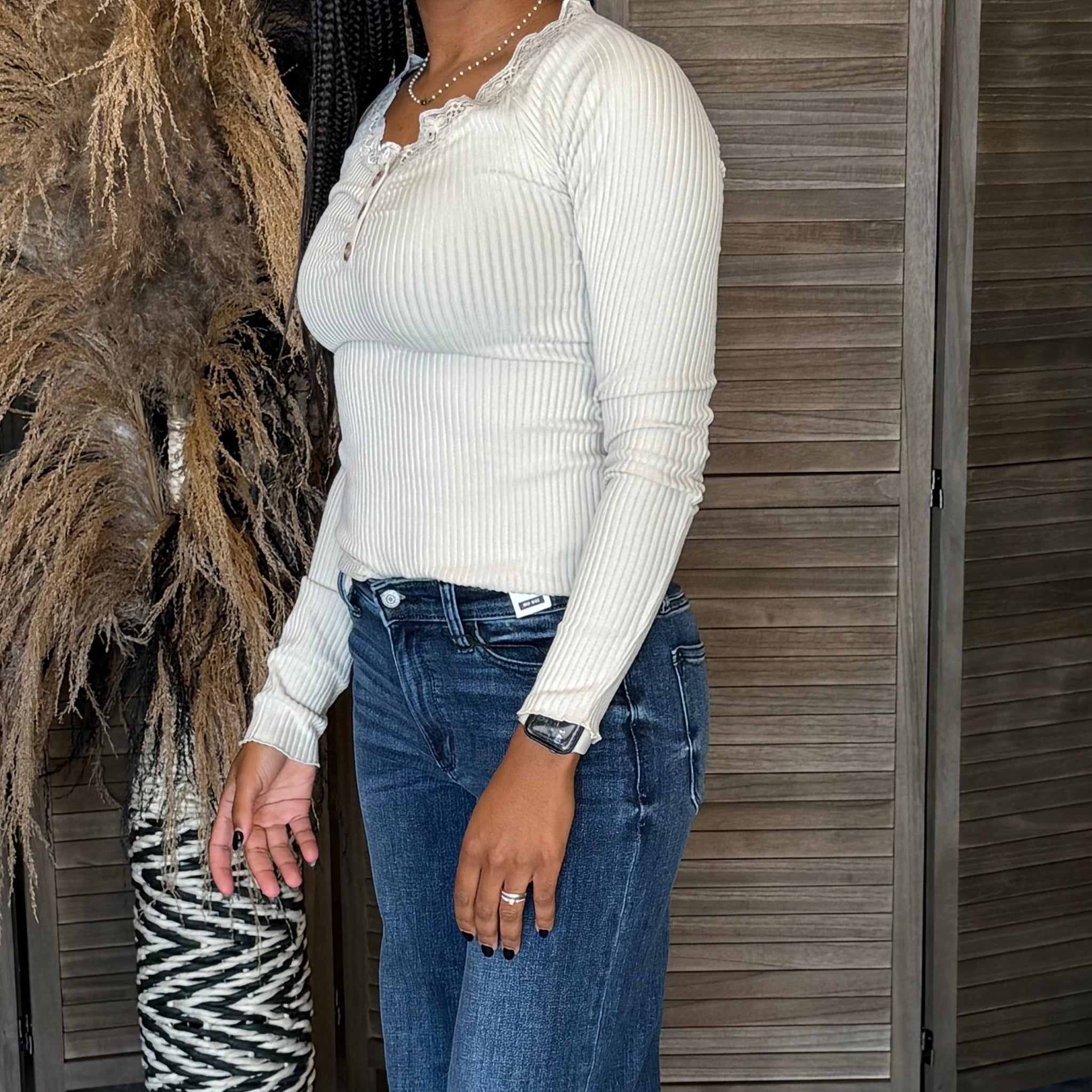 Person wearing a white ribbed long-sleeve top and blue jeans standing against a wooden wall.