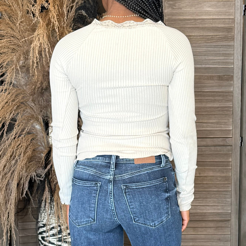 Person wearing a white ribbed sweater and blue jeans standing in front of a wooden wall.