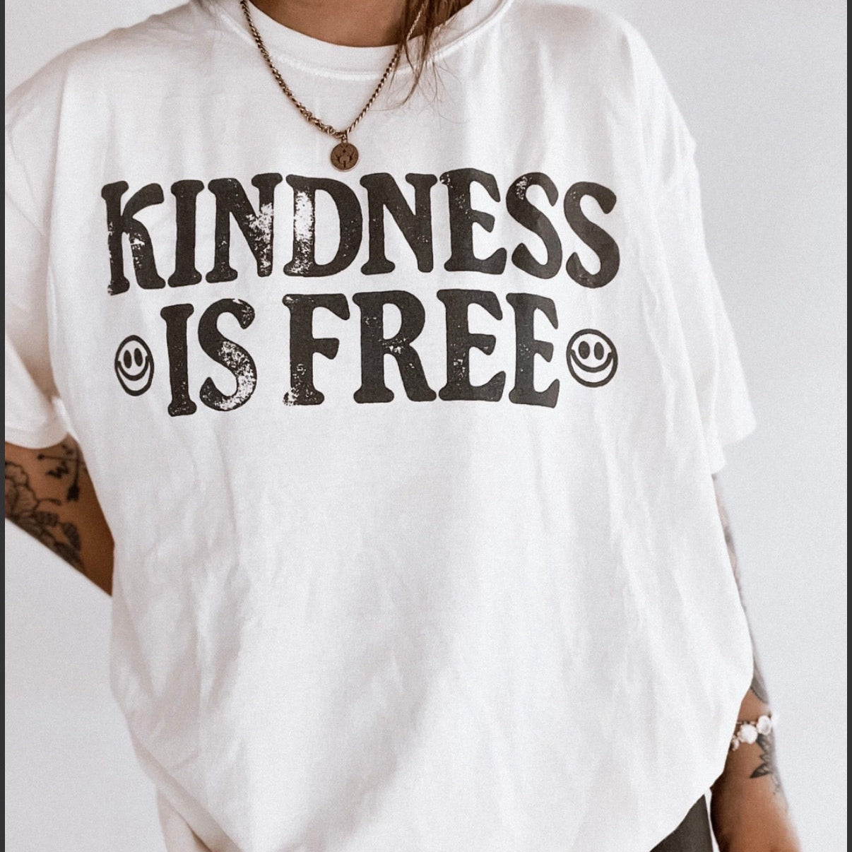 White t-shirt with 'Kindness is Free' text and smiley faces on a plain background