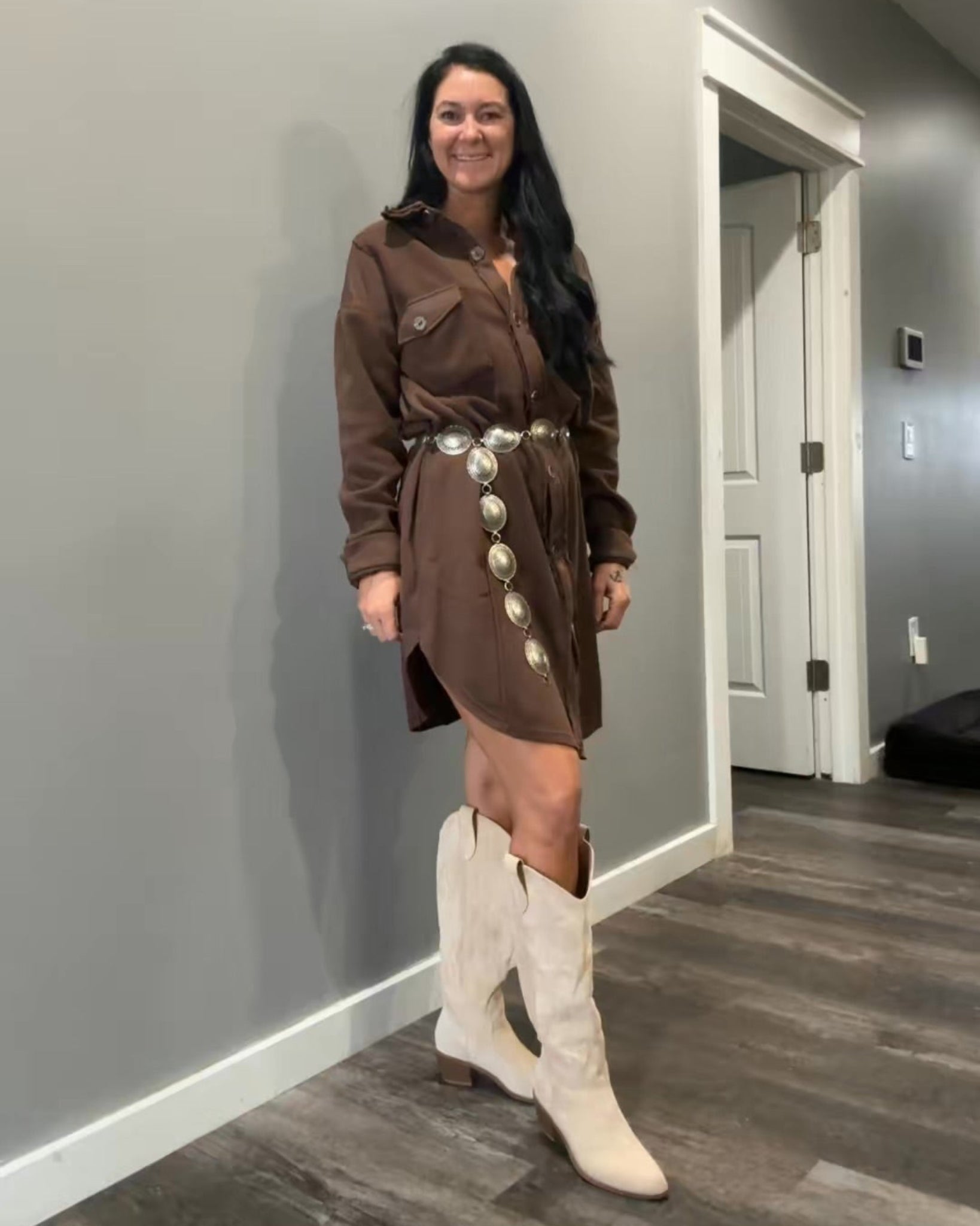 Woman in a brown dress with white buttons and white boots standing in a room with gray walls and wooden floor.