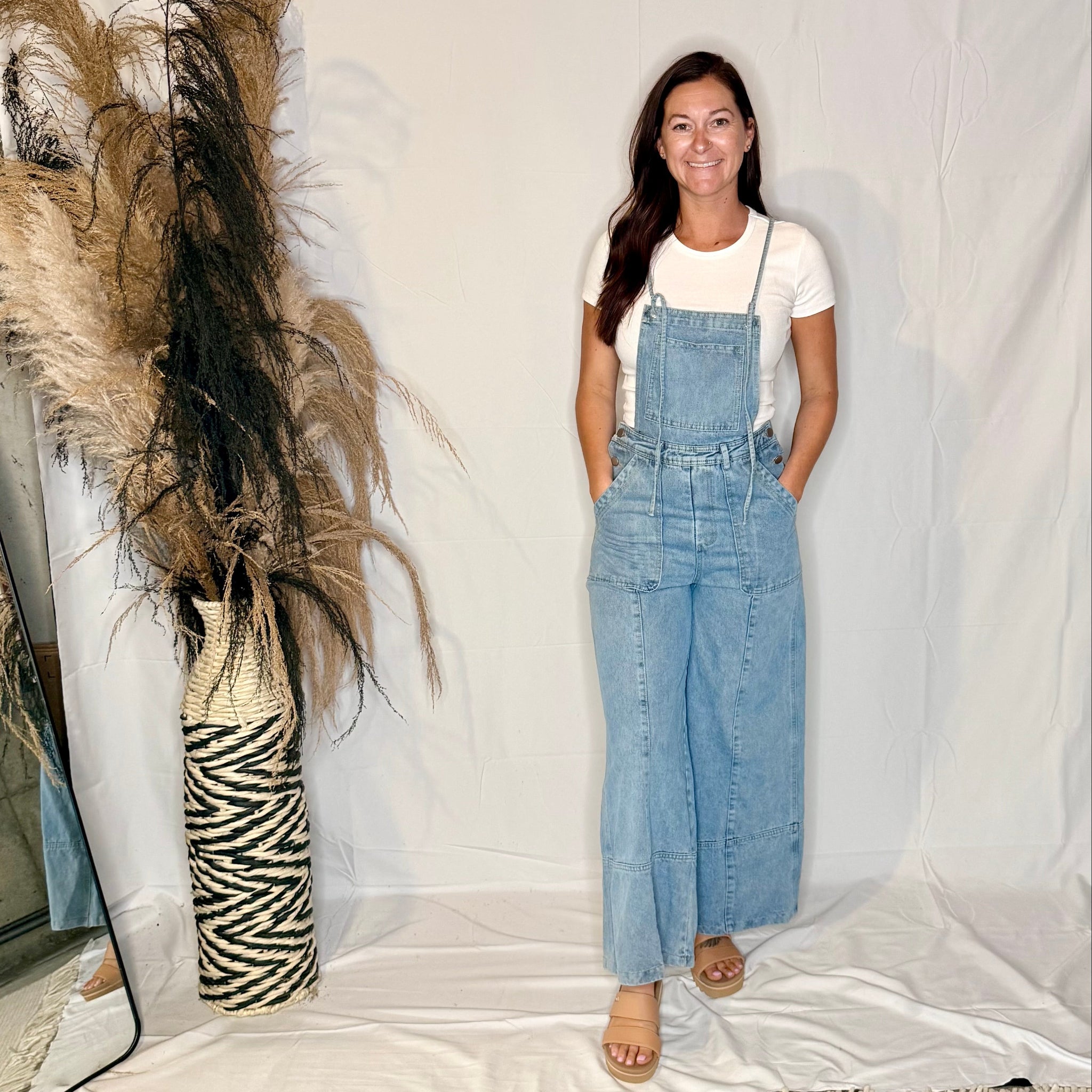 Woman wearing a denim jumpsuit standing next to decorative pampas grass and a zebra-patterned vase on a white background.