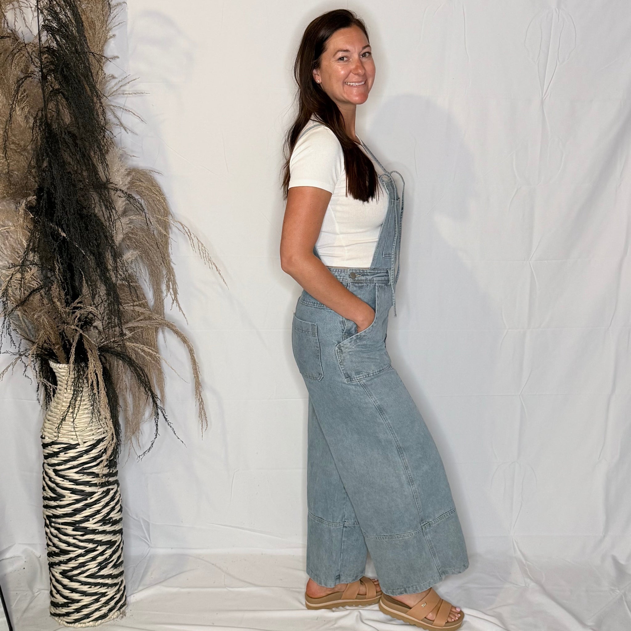 Woman wearing denim overalls standing next to a decorative plant against a white backdrop