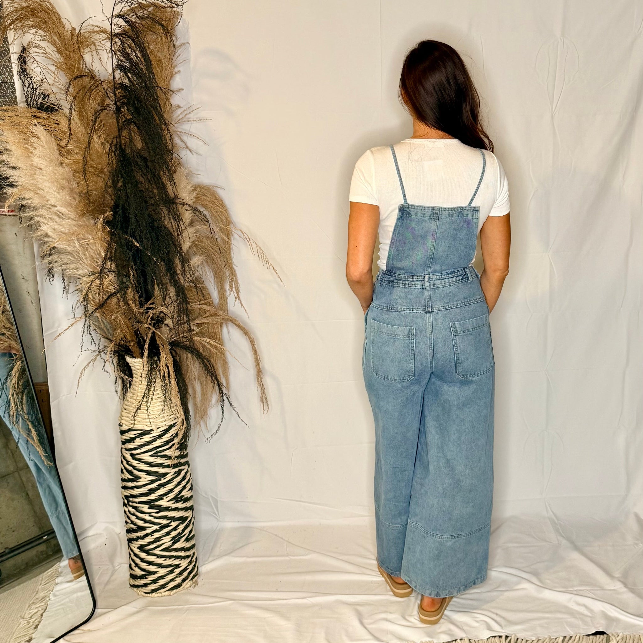 Person wearing a denim overalls standing in front of a decorative vase with pampas grass on a white background