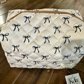 A quilted white cosmetic bag with a repeating black bow pattern and a gold zipper, resting on a dark wood floor with a Kyla Rae tag attached.