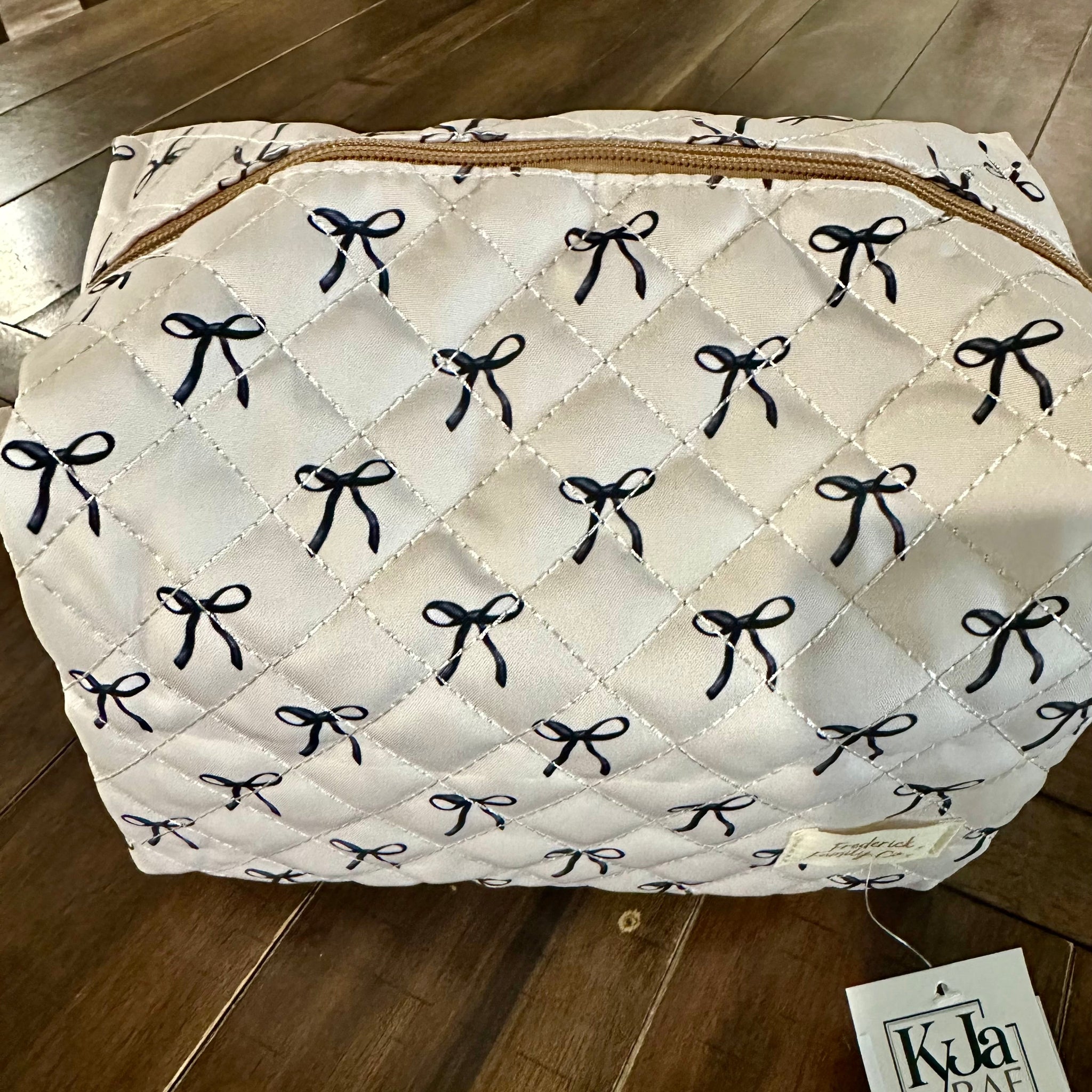 A quilted white cosmetic bag with a repeating black bow pattern and a gold zipper, resting on a dark wood floor with a Kyla Rae tag attached.
