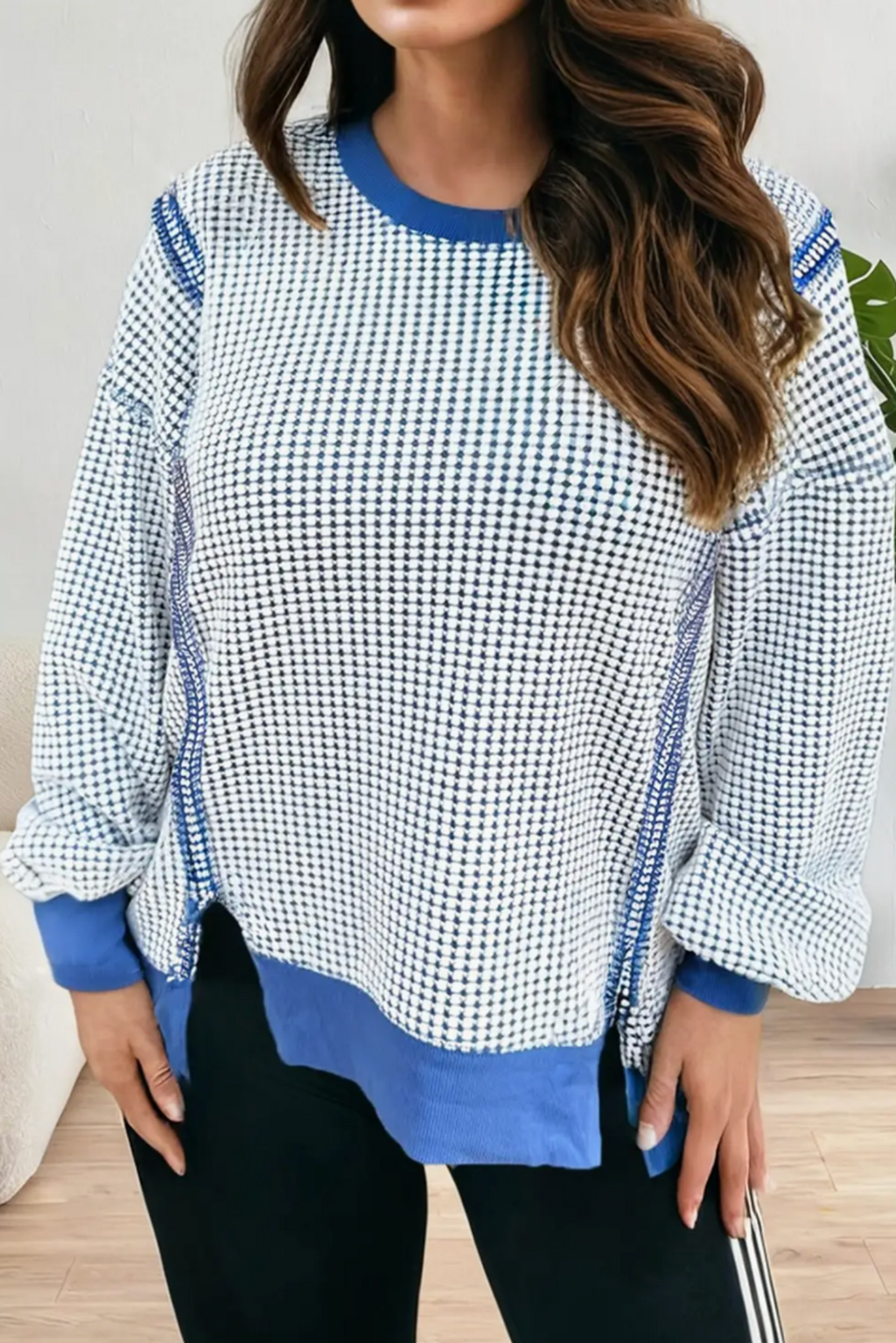A woman wearing a long-sleeve crewneck top with a blue and white micro-check pattern, solid blue ribbed collar, cuffs, and hem, paired with black pants.
