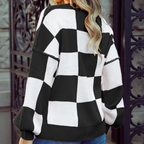 A woman wearing a black and white checkered knit sweater and blue jeans, photographed from the back and side against a stone wall and dark metal gate.