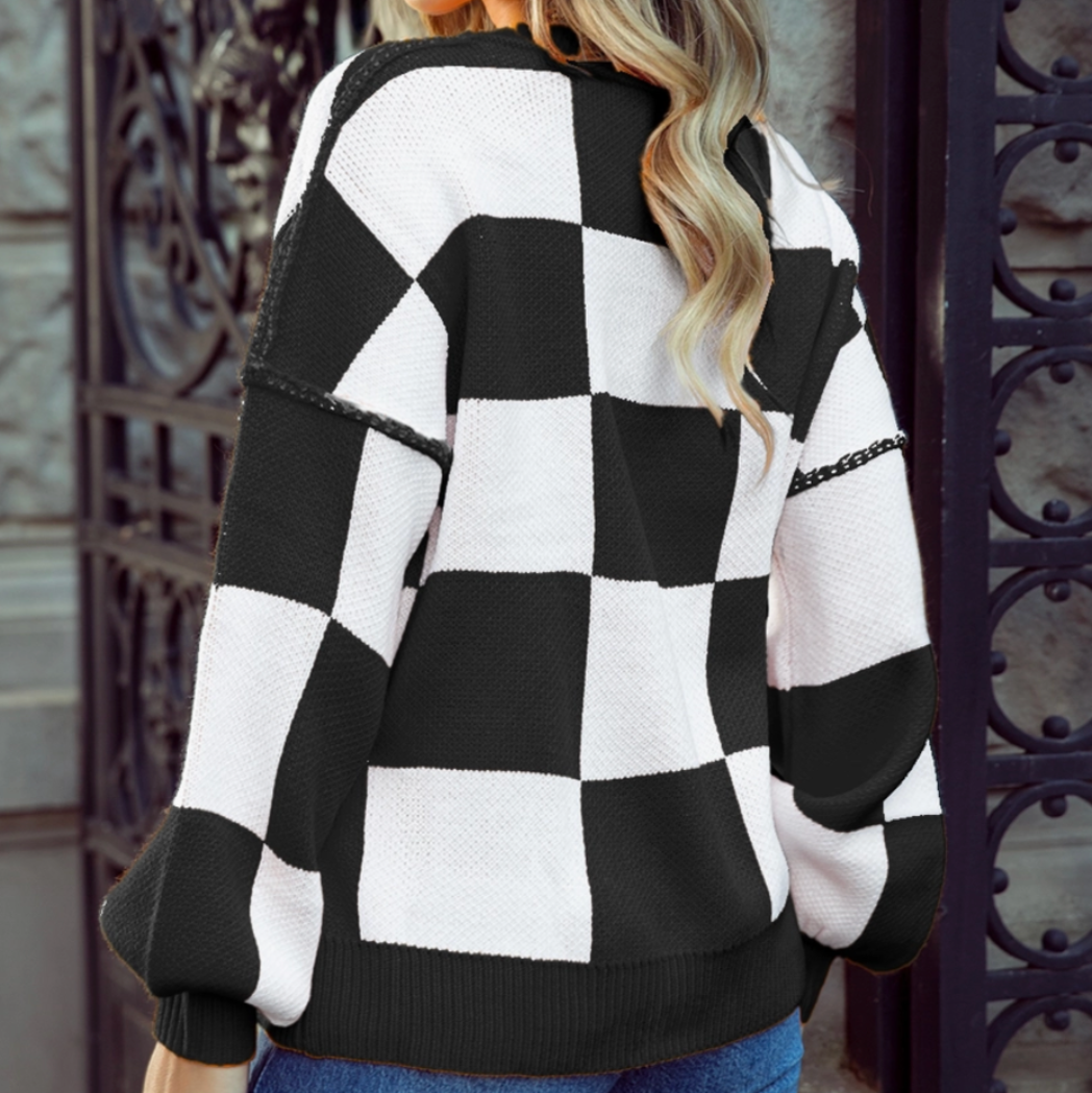 A woman wearing a black and white checkered knit sweater and blue jeans, photographed from the back and side against a stone wall and dark metal gate.