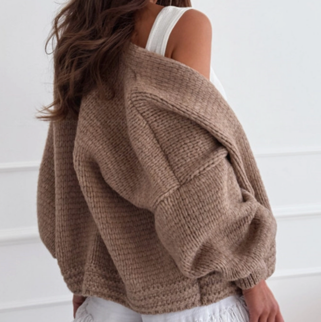 A woman wearing an oversized chunky knit brown sweater draped off her shoulders, paired with a white tank top and white fringed shorts, photographed against a white paneled wall.