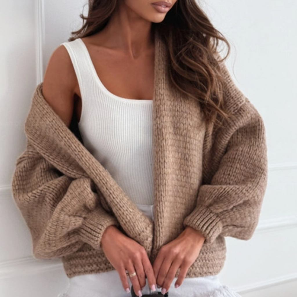 A woman wearing a chunky taupe knit cardigan over a white ribbed tank top and white fringed shorts, holding a brown Louis Vuitton monogram handbag against a white paneled wall.