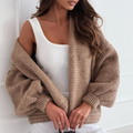 A woman wearing a chunky taupe knit cardigan over a white ribbed tank top and white fringed shorts, holding a brown Louis Vuitton monogram handbag against a white paneled wall.