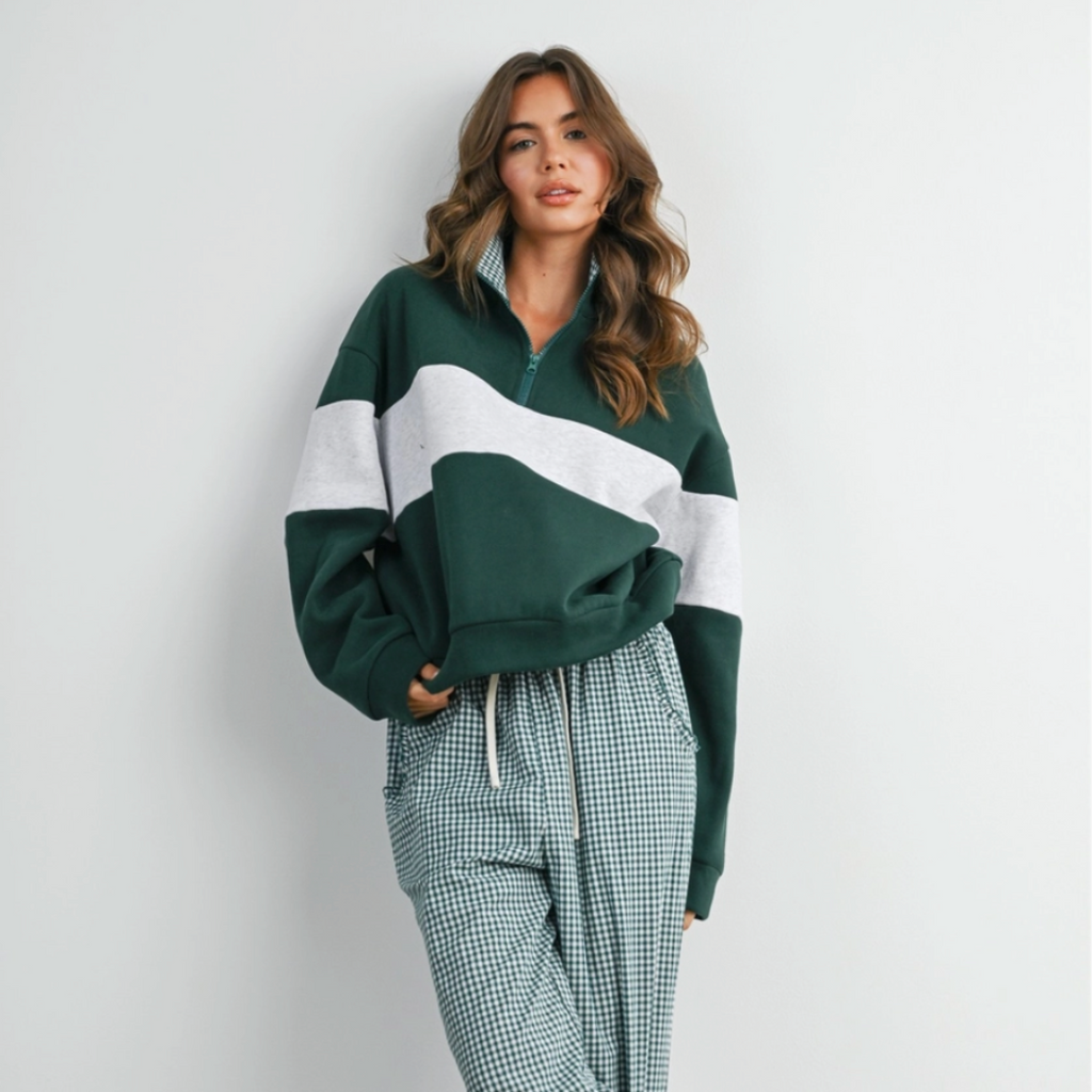 A woman modeling a cozy matching loungewear set consisting of a dark green and white color-blocked quarter-zip sweatshirt and matching green-and-white gingham wide-leg pants, paired with brown slip-on shoes against a plain white wall.