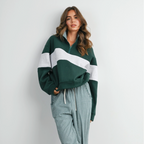 A woman modeling a cozy matching loungewear set consisting of a dark green and white color-blocked quarter-zip sweatshirt and matching green-and-white gingham wide-leg pants, paired with brown slip-on shoes against a plain white wall.