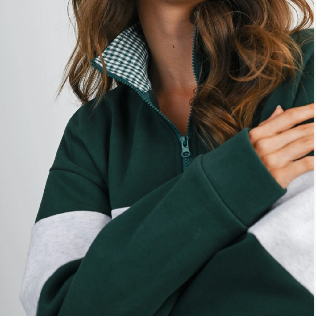 A woman wearing a dark green color-blocked sweatshirt with light gray sleeves and a green-and-white gingham check collar, photographed against a plain light gray background.