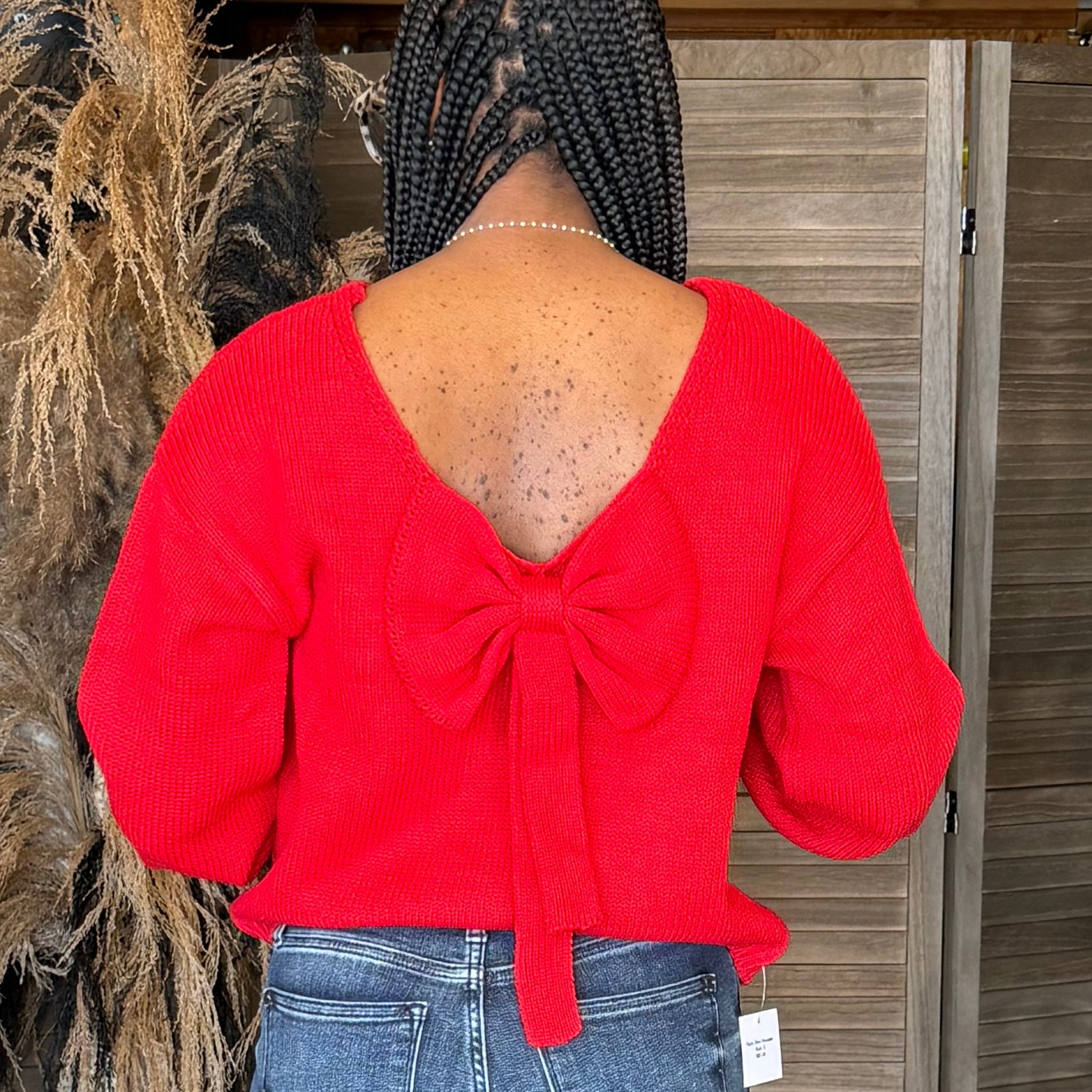 Person wearing a red sweater with a bow detail and blue jeans, standing against a wooden wall.