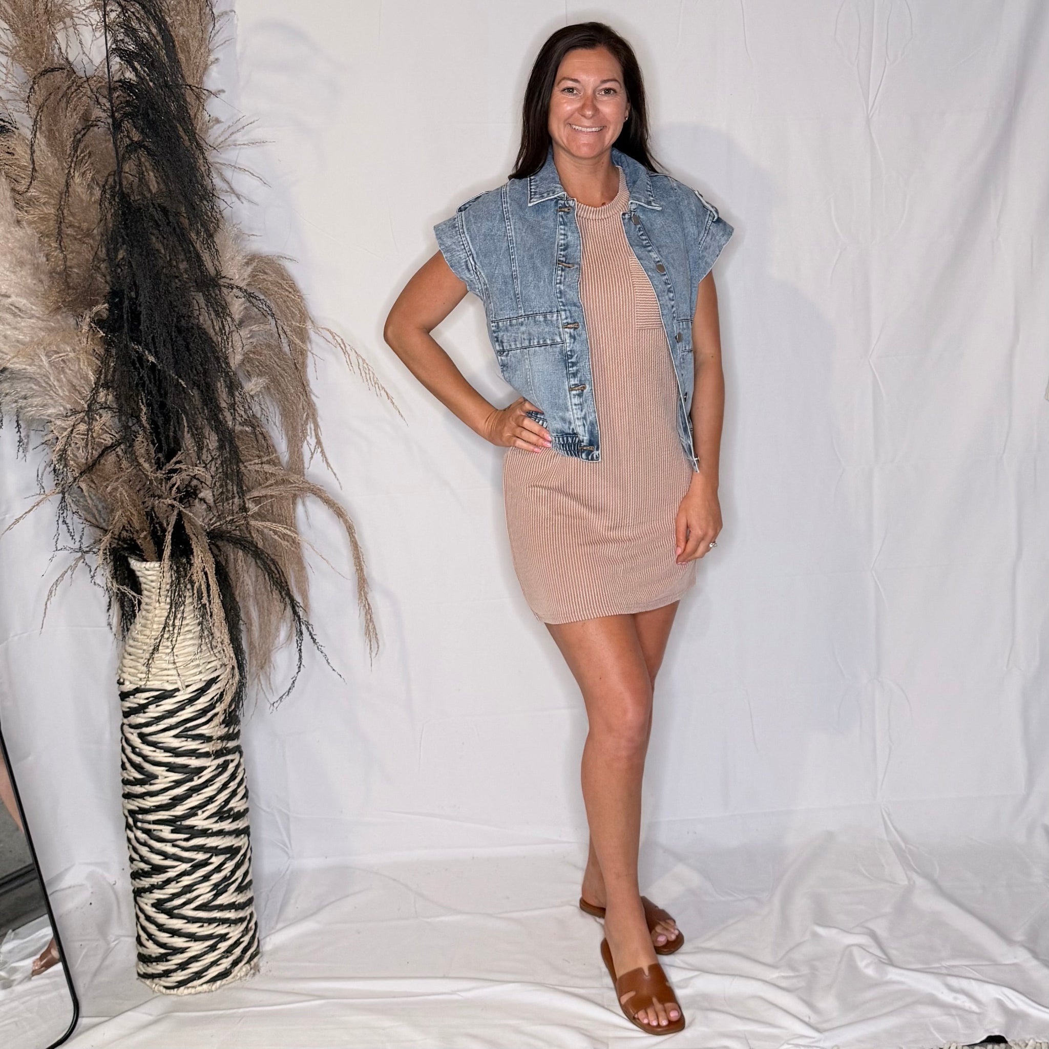 Woman wearing a beige dress with a denim vest standing next to a decorative vase against a white background