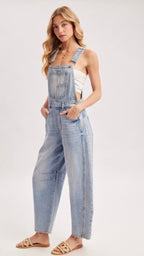 A woman modeling a pair of light-wash blue denim overall jumpsuit with wide legs, styled with a white tank top and tan slide sandals against a plain light gray studio background.