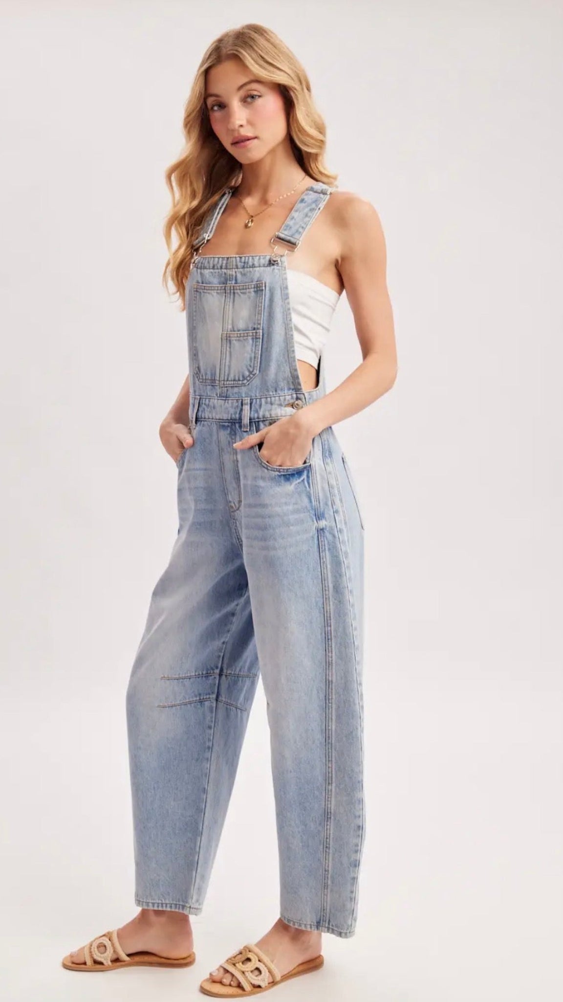 A woman modeling a pair of light-wash blue denim overall jumpsuit with wide legs, styled with a white tank top and tan slide sandals against a plain light gray studio background.