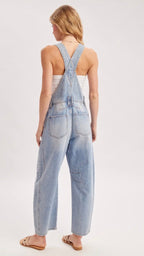 A woman viewed from behind wearing light-wash blue denim overall with wide legs and crossed shoulder straps, paired with a white tank top and tan sandals, against a plain light gray studio background.