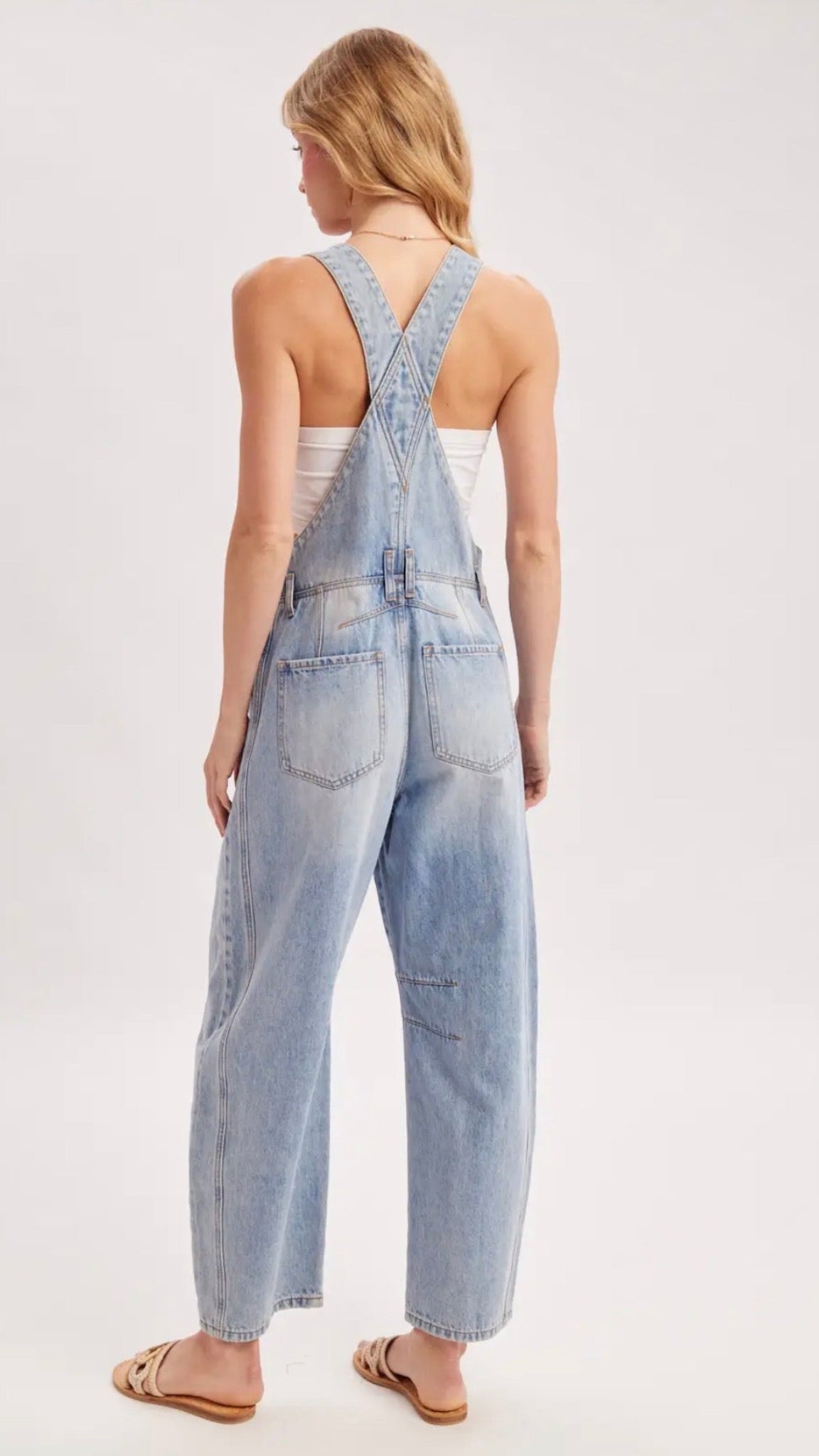 A woman viewed from behind wearing light-wash blue denim overall with wide legs and crossed shoulder straps, paired with a white tank top and tan sandals, against a plain light gray studio background.