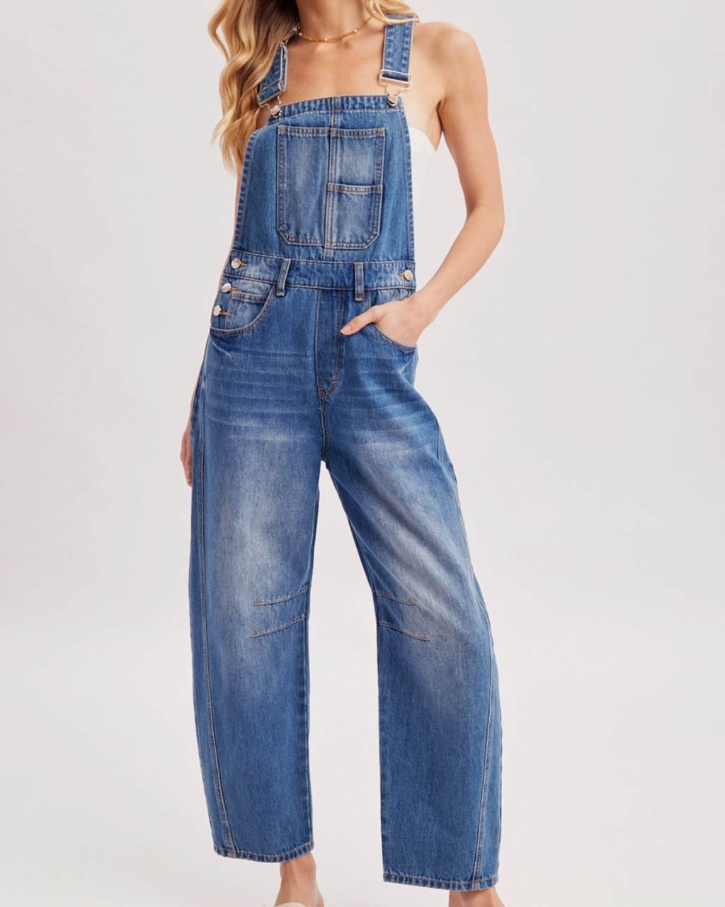 A woman modeling a pair of wide-leg blue denim overalls with front patch pockets and adjustable shoulder straps, styled with a brown wide-brim hat and beige slide sandals against a light gray studio background.