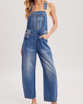 A woman modeling a pair of wide-leg blue denim overalls with front patch pockets and adjustable shoulder straps, styled with a brown wide-brim hat and beige slide sandals against a light gray studio background.