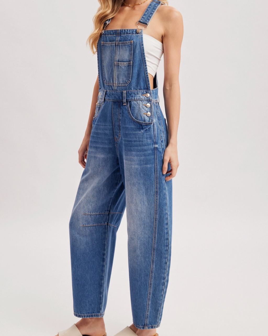 A woman modeling a pair of wide-leg blue denim overalls with a white tank top, a brown wide-brim hat, and beige slide sandals against a light gray studio background.