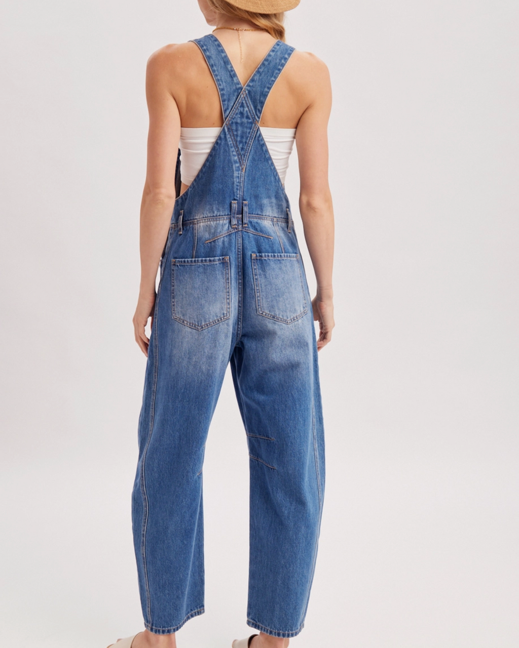 A model shown from the back wearing blue denim overall-style shorts with wide legs, a tan wide-brim hat, a white tank top, and tan flat sandals, photographed against a clean light gray studio background.
