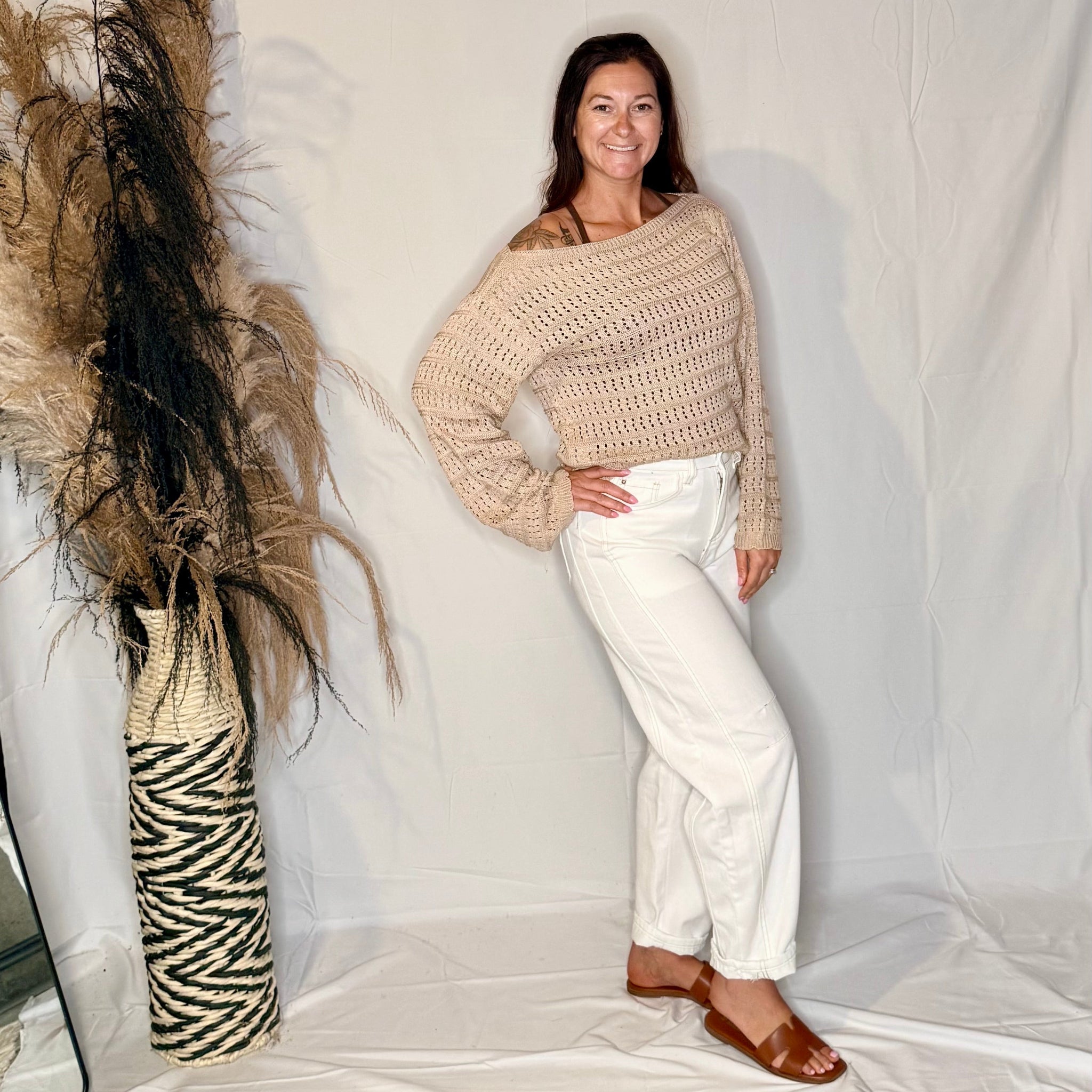 Woman wearing a beige sweater and white pants standing next to decorative plants against a plain background