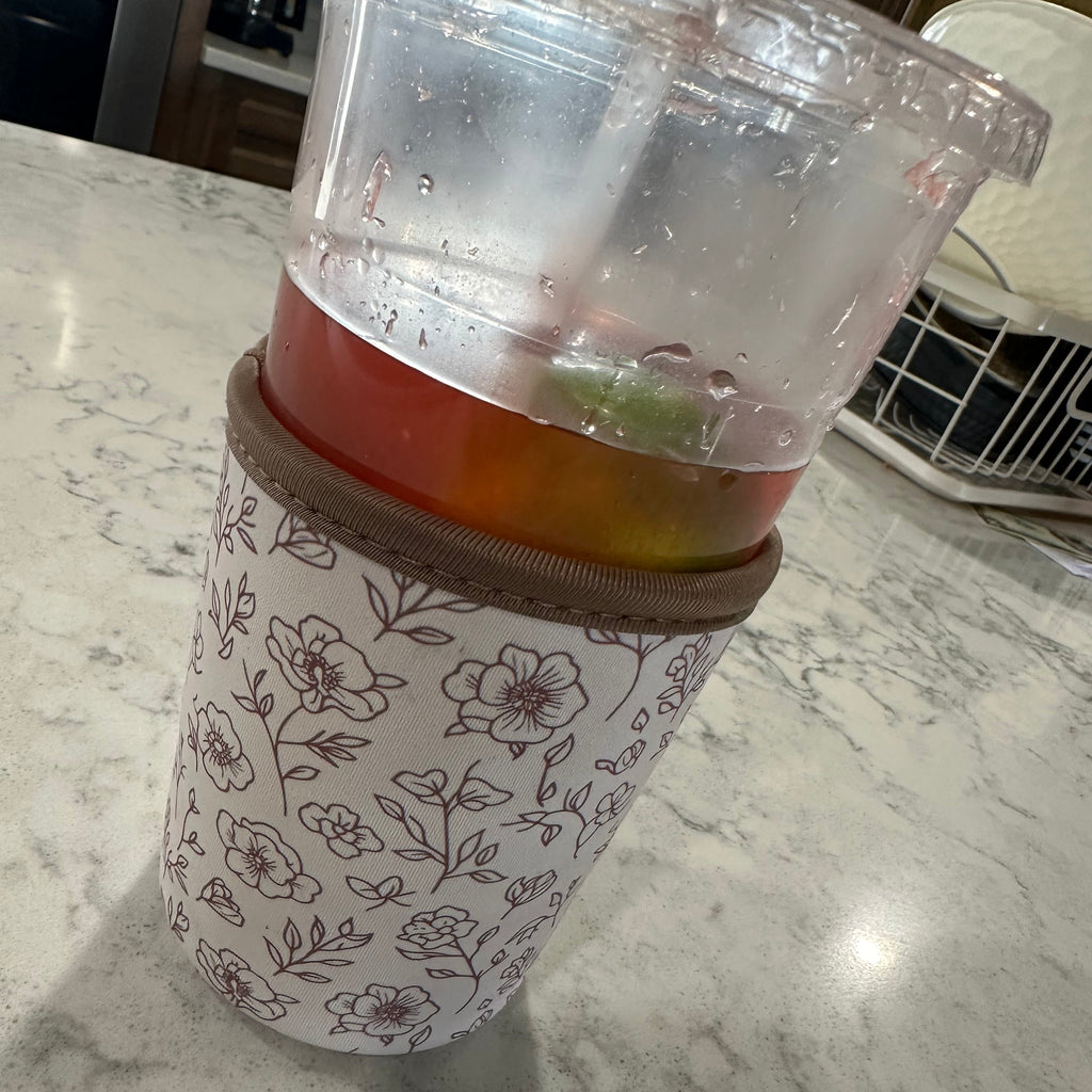 A clear plastic travel tumbler with a brown lid and a white sleeve printed with a brown floral pattern, resting on a white marble countertop.
