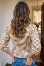 A woman viewed from behind wearing a brown and beige gingham check long-sleeve peplum top and blue jeans, standing in a doorway.