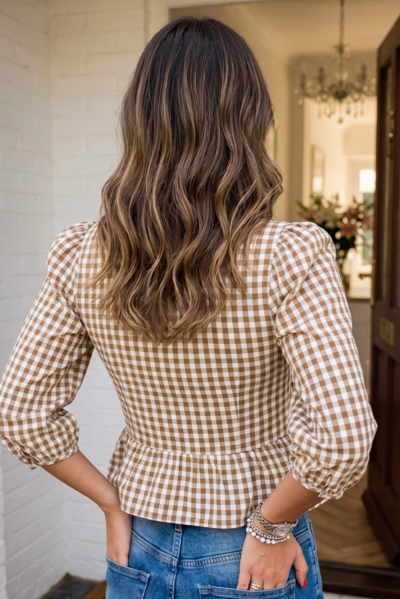 A woman viewed from behind wearing a brown and beige gingham check long-sleeve peplum top and blue jeans, standing in a doorway.