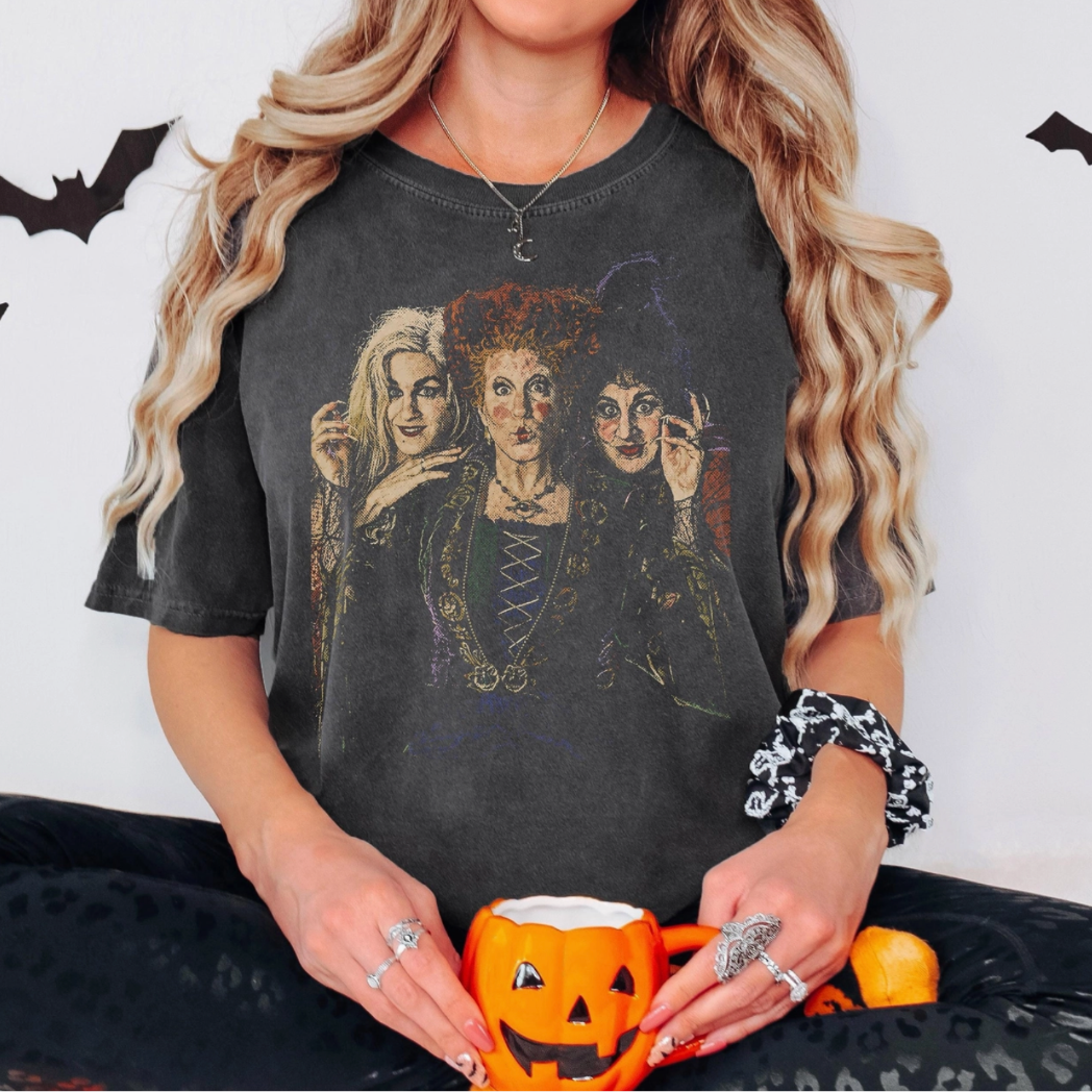 A woman wearing a faded black graphic t-shirt featuring the characters from 'The Addams Family' in a Halloween-themed setting, holding a jack-o'-lantern mug.