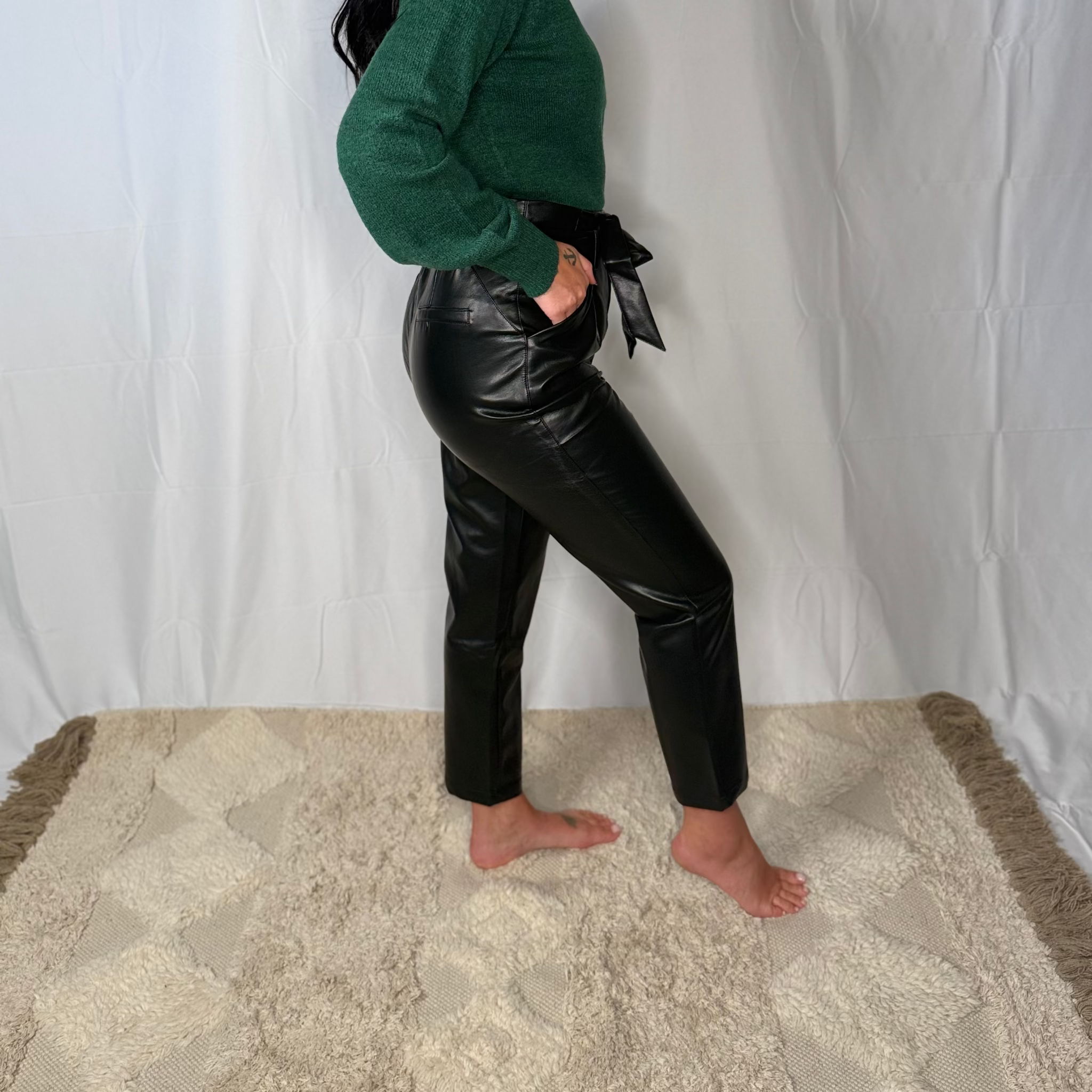 Woman wearing a green sweater and black leather pants standing against a white curtain.