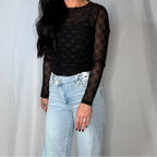 Person wearing a black sheer top with long sleeves and light blue jeans against a plain background