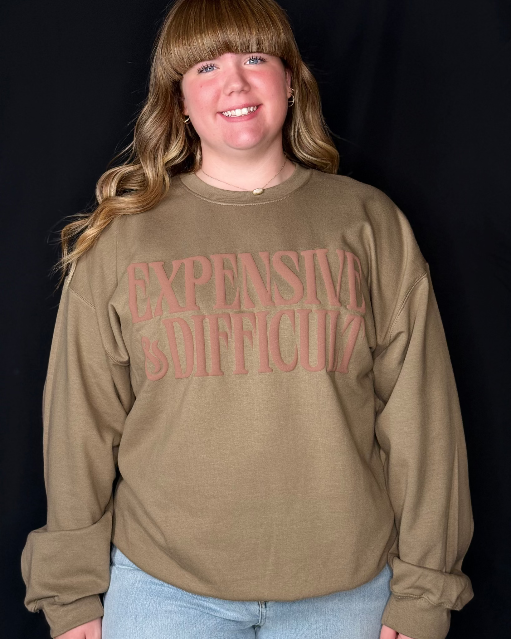 Person wearing a brown sweatshirt with 'EXPENSIVE IS DIFFICULT' text on a black background