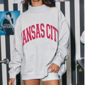 A woman wearing an oversized light gray sweatshirt with 'KANSAS CITY' printed in red across the chest, standing in front of a black-and-white striped wall with posters.