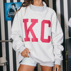 A woman wearing a white crewneck sweatshirt with large pink block letters 'KC' on the front, paired with light blue and white patterned shorts, standing in front of a black and white striped wall with posters.