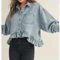 A woman wearing a light-wash blue denim shirt jacket with a ruffled hem and voluminous sleeves, paired with black pants, against a plain beige background.