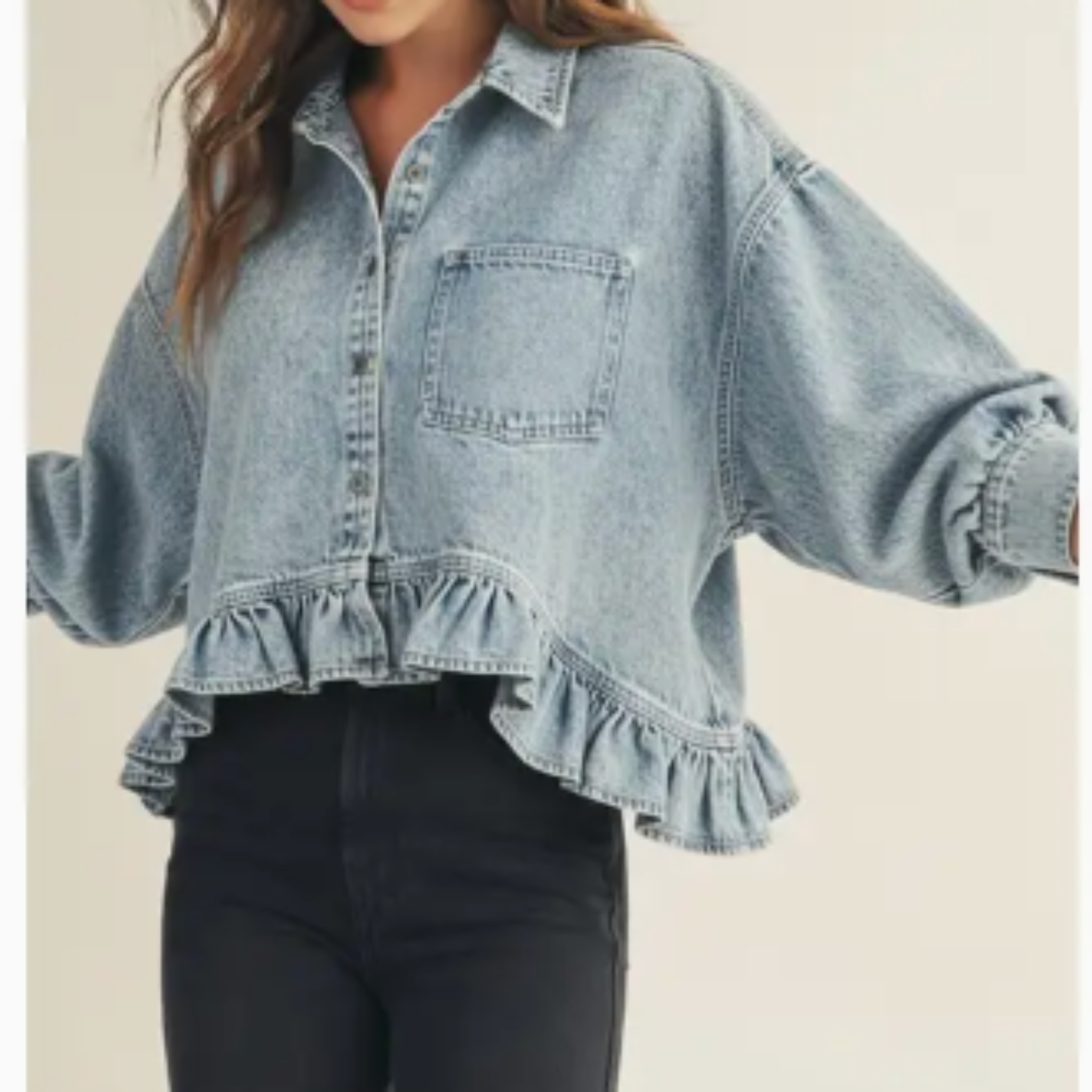 A woman wearing a light-wash blue denim shirt jacket with a ruffled hem and voluminous sleeves, paired with black pants, against a plain beige background.