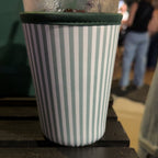 A clear glass tumbler with a dark green ribbed sleeve, featuring vertical white and light blue-gray stripes, sitting on a dark wooden table in a dimly lit indoor setting.