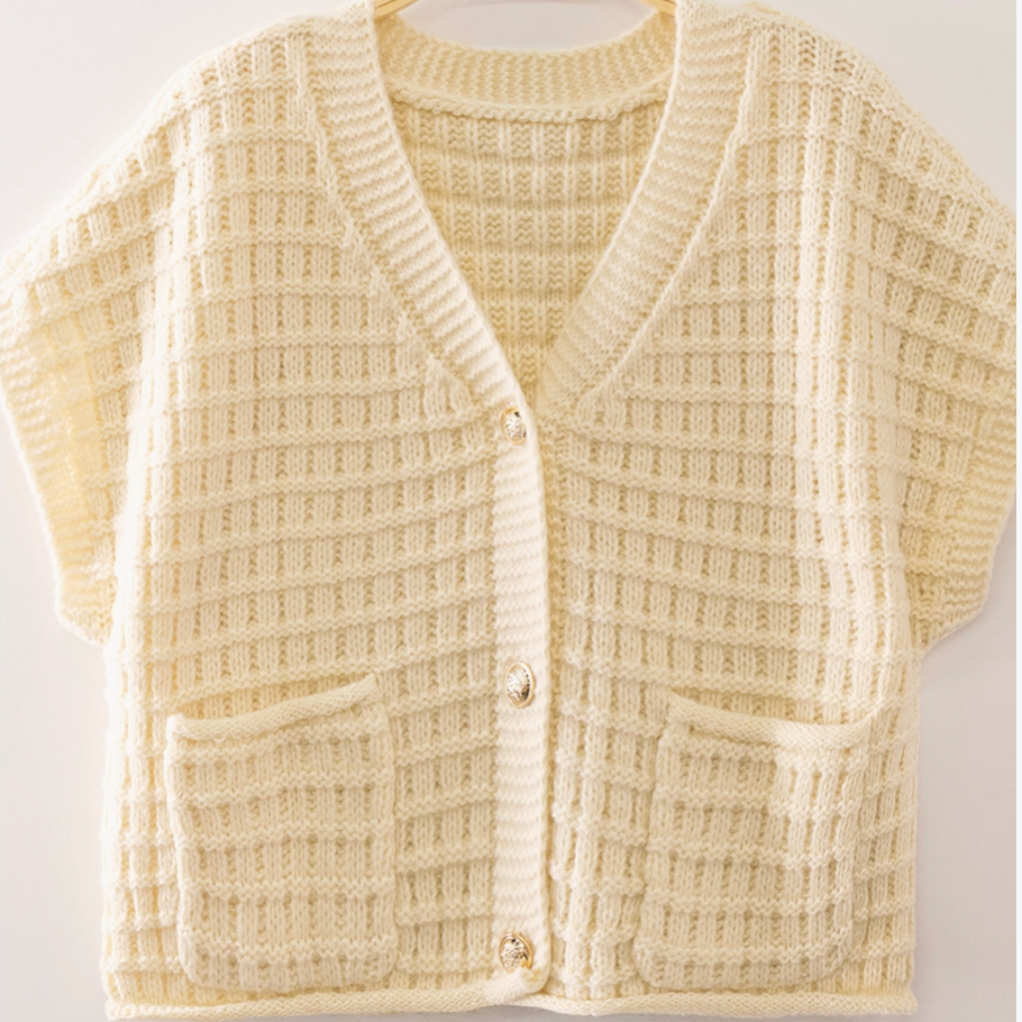 A cream-colored waffle-knit cardigan sweater with short sleeves, a V-neck, and two front patch pockets, hanging on a wooden hanger against a light background.