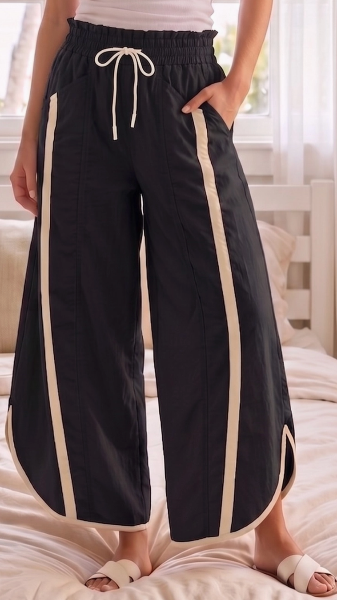 A person wearing black wide-leg pants with cream-colored side stripes and a cream drawstring waist, standing on a bed in a bedroom setting.
