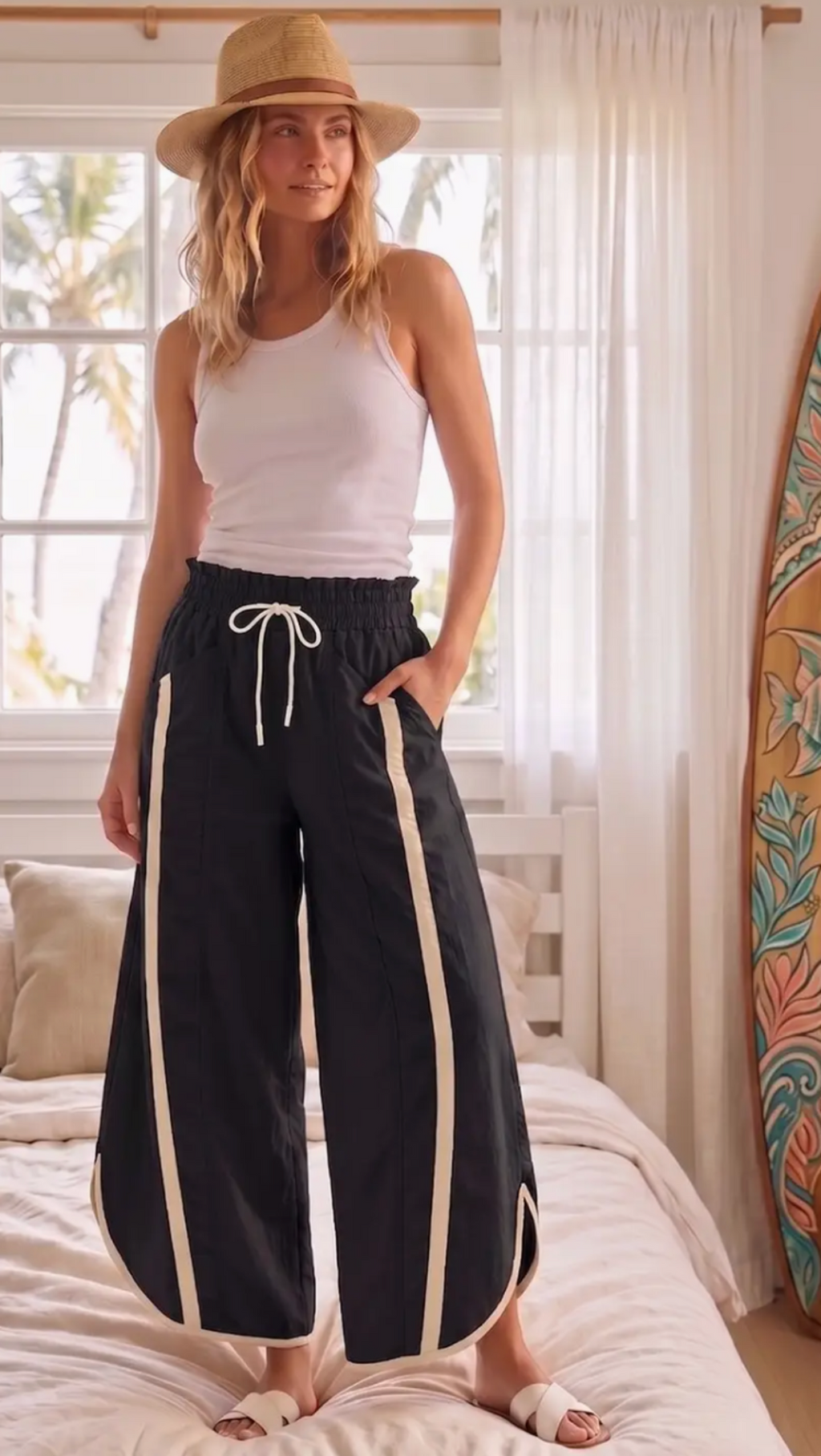A woman stands on a bed wearing loose black wide-leg pants with cream side stripes and a white tank top, paired with a tan hat and white slide sandals. The room has a window with white curtains and a surfboard leaning against the wall.
